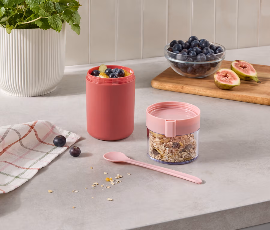 Deux récipients sur un comptoir de cuisine: un grand récipient rose rempli de fruits et un récipient transparent avec du muesli et un couvercle rose. À côté, il y a une cuillère rose, un torchon à carreaux, des baies, un bol de myrtilles et des figues coupées sur une planche en bois.