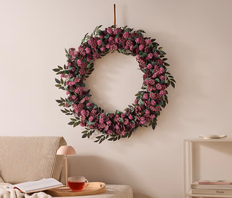 Grande couronne florale de fleurs violettes et de feuilles vertes accrochée à un mur clair. À gauche, un canapé avec une couverture, un livre, une lampe rose et une tasse de thé sur un plateau. À droite, une étagère blanche avec des livres.