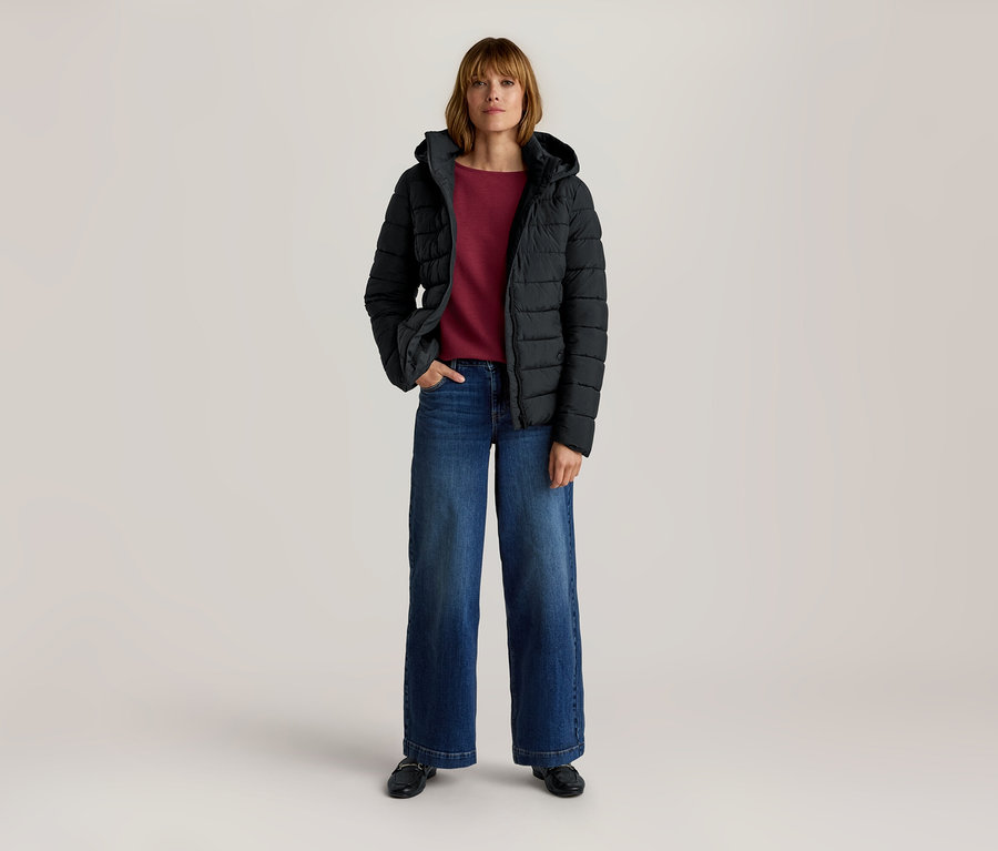 Femme debout portant une doudoune noire à capuche, un haut rouge à manches longues, un jean large bleu et des mocassins noirs, une main dans la poche.