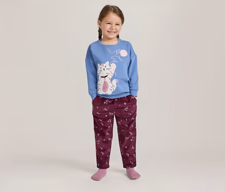 Fille souriante avec des tresses, portant un pull bleu à manches longues avec un motif de chat et un pantalon bordeaux à motifs.