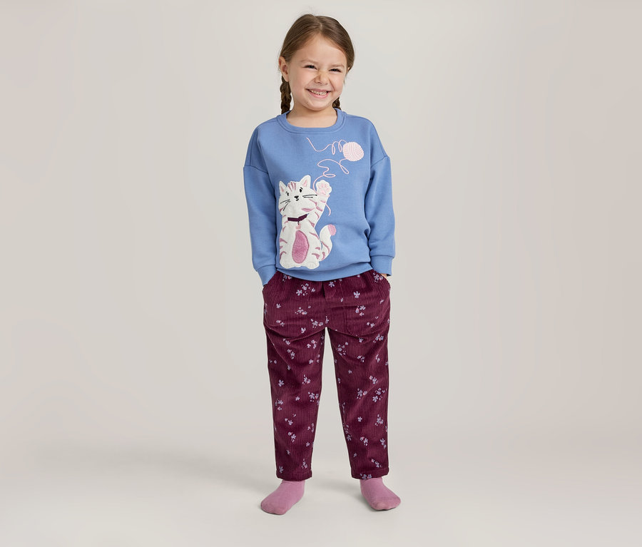 Fille souriante avec des tresses, portant un pull bleu à manches longues avec un motif de chat et un pantalon bordeaux à motifs.