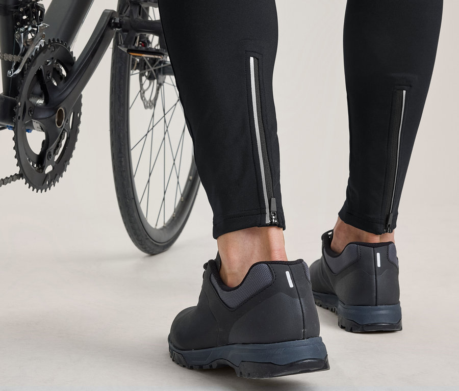 Jambes et pieds d'une personne debout à côté d'un vélo. Elle porte un pantalon noir avec des fermetures éclair réfléchissantes aux chevilles et des chaussures de cyclisme noires.
