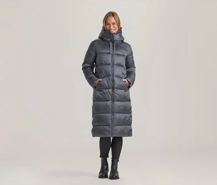 Femme souriante debout, vêtue d'un long manteau doudoune gris, les mains dans les poches, avec un legging noir et des bottes noires.