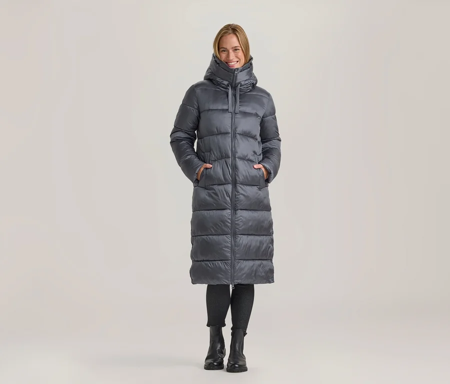 Femme souriante debout, vêtue d'un long manteau doudoune gris, les mains dans les poches, avec un legging noir et des bottes noires.