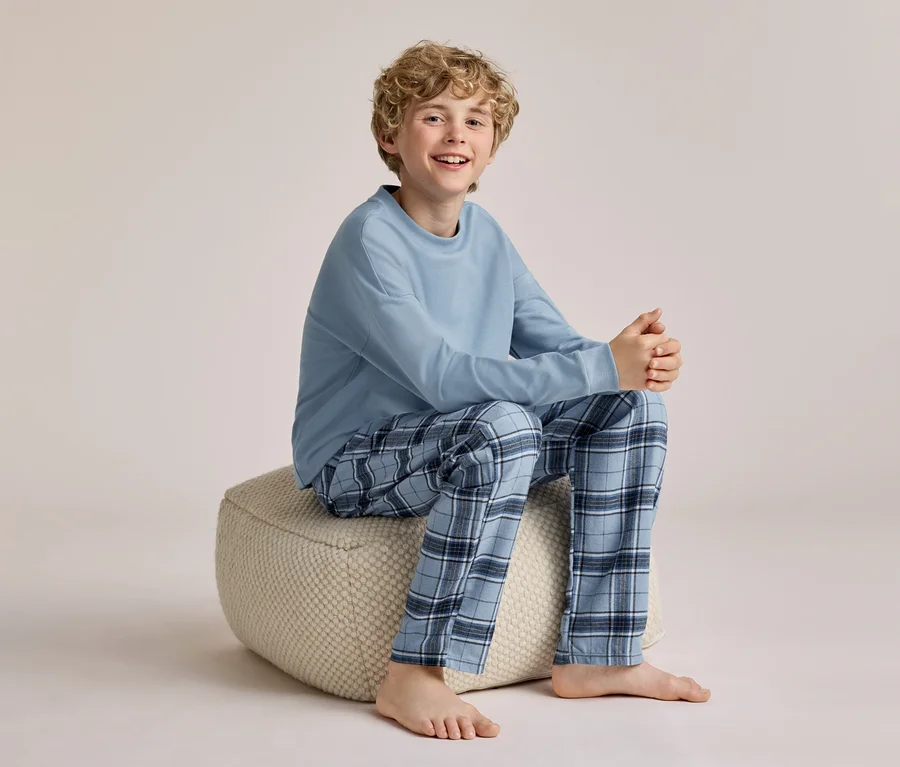 Un jeune garçon souriant aux cheveux blonds bouclés est assis pieds nus sur un pouf clair, portant un haut bleu clair à manches longues et un pantalon de pyjama à carreaux bleus.