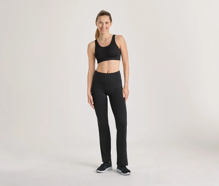 Femme souriante debout, vêtue d'un soutien-gorge de sport noir, d'un pantalon de sport noir et de baskets noires.