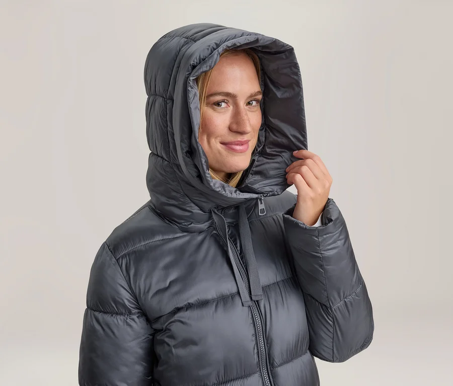 Femme souriante portant une doudoune gris foncé à capuche, qu'elle tient d'une main.