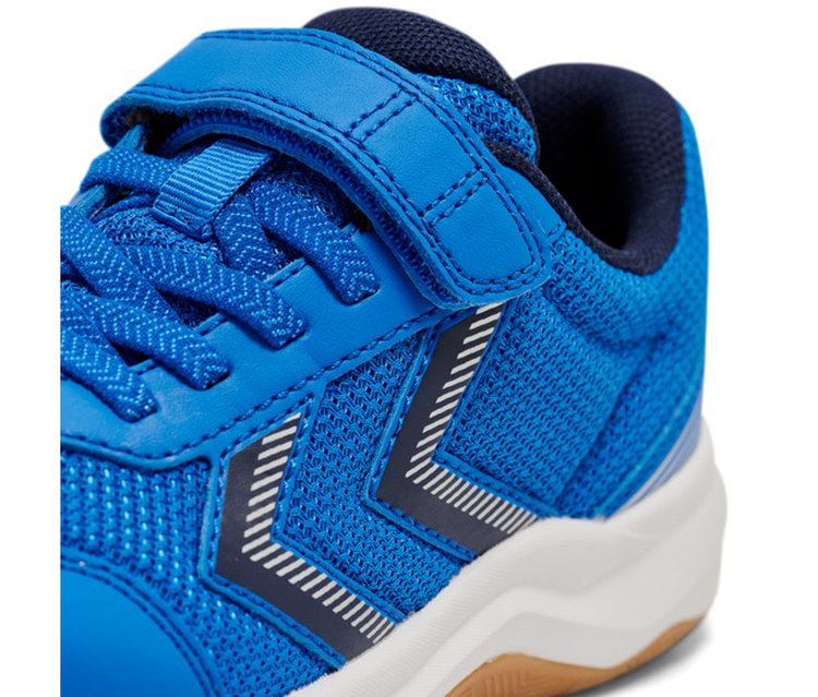 Gros plan d'une basket bleue avec des détails en maille et en cuir synthétique. Elle présente des lacets bleus, une sangle Velcro bleue et un logo rayé blanc et bleu foncé sur le côté. La semelle est blanche avec une semelle extérieure marron.