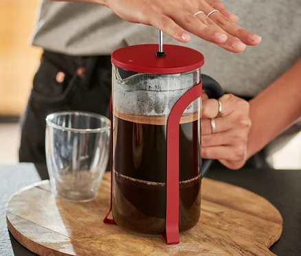 Femme appuyant sur une cafetière à piston rouge de 800 ml, 6 tasses sur une planche à découper en bois.