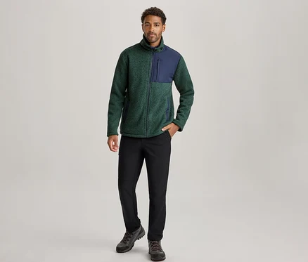 Homme debout, portant une veste polaire verte et bleu marine, un pantalon noir et des chaussures de randonnée foncées, la main gauche dans la poche.