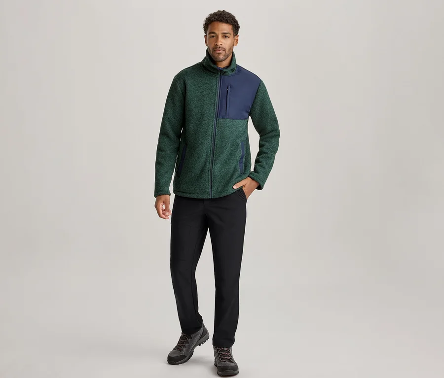 Homme debout, portant une veste polaire verte et bleu marine, un pantalon noir et des chaussures de randonnée foncées, la main gauche dans la poche.