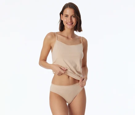 Femme souriante portant un débardeur à fines bretelles et une culotte beiges, soulevant légèrement son débardeur.