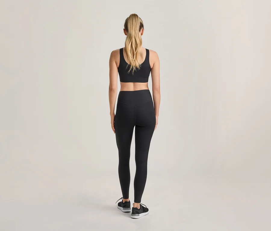 Femme vue de dos, portant un soutien-gorge de sport noir, un legging noir et des chaussures de sport noires, avec ses cheveux blonds attachés en queue de cheval.