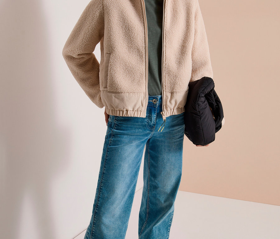 Une femme souriante porte une veste CECIL beige à capuche, un t-shirt vert, un jean bleu et des baskets blanches. Elle tient un sac noir.
