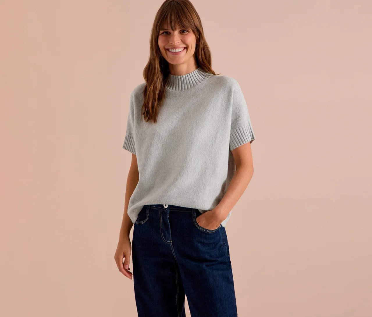 Femme souriante aux longs cheveux bruns, portant un pull en maille CECIL gris à manches courtes et un jean foncé.