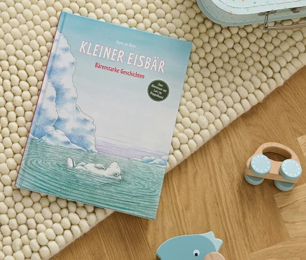 Le livre «	Kleiner Eisbär – bärenstarke Geschichten	» posé sur un tapis beige à côté de jouets en bois.