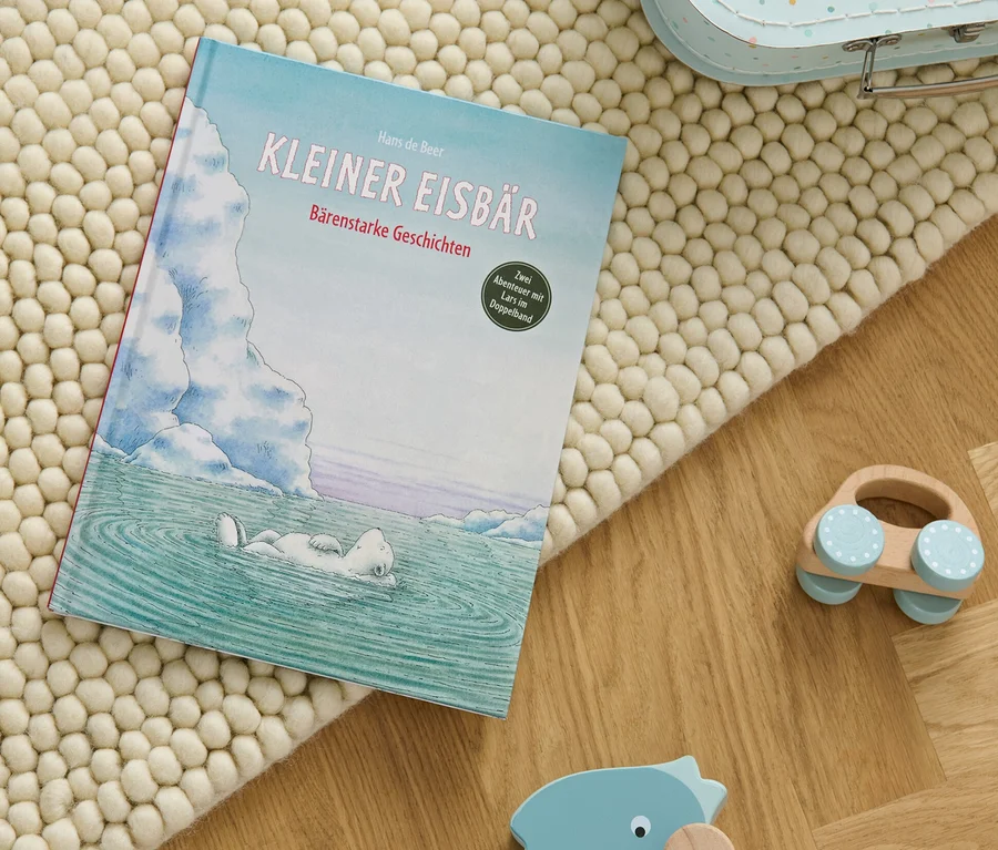 Le livre «	Kleiner Eisbär – bärenstarke Geschichten	» posé sur un tapis beige à côté de jouets en bois.