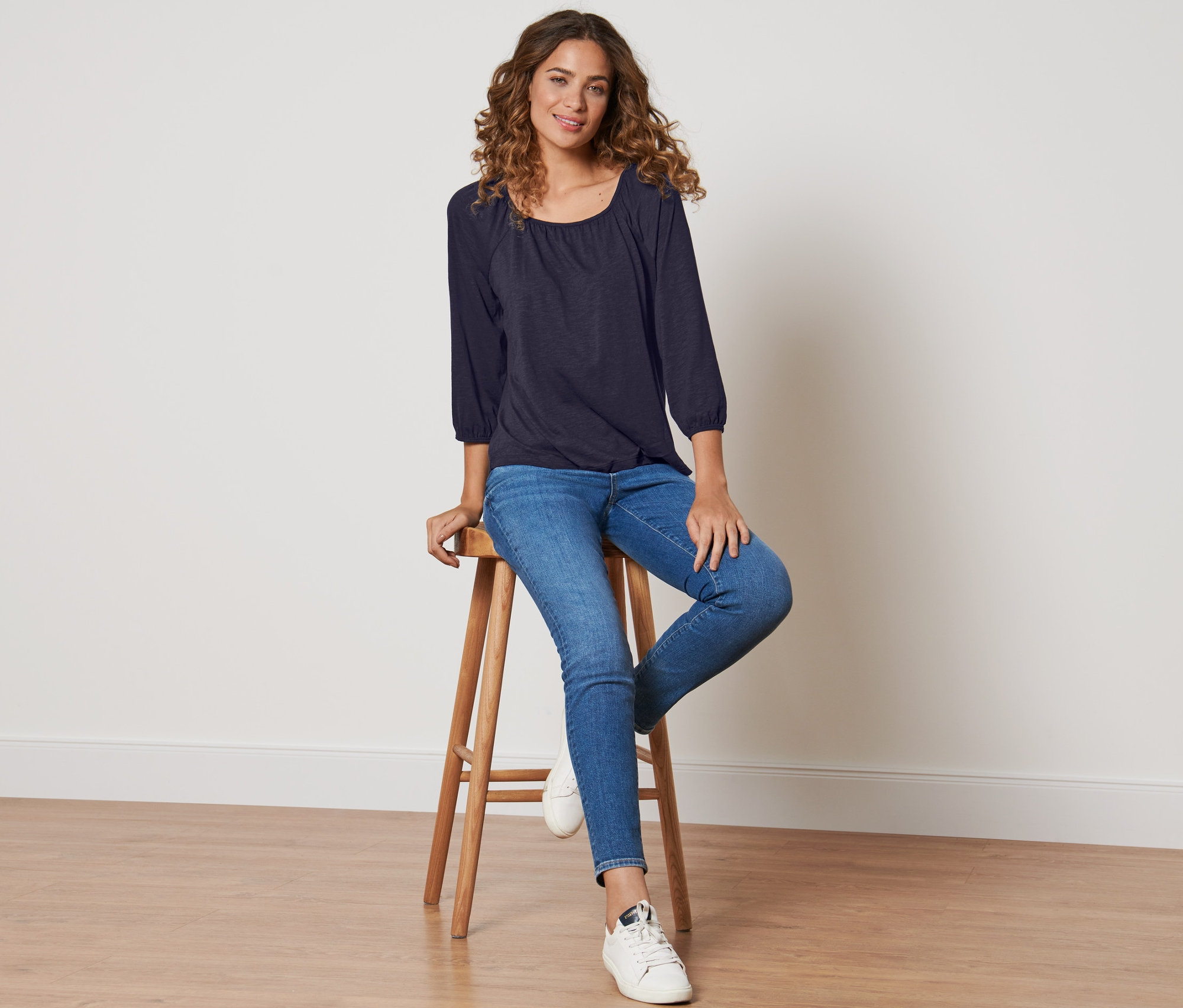 Une femme assise sur un tabouret en bois portant une blouse bleu foncé à manches 3/4.