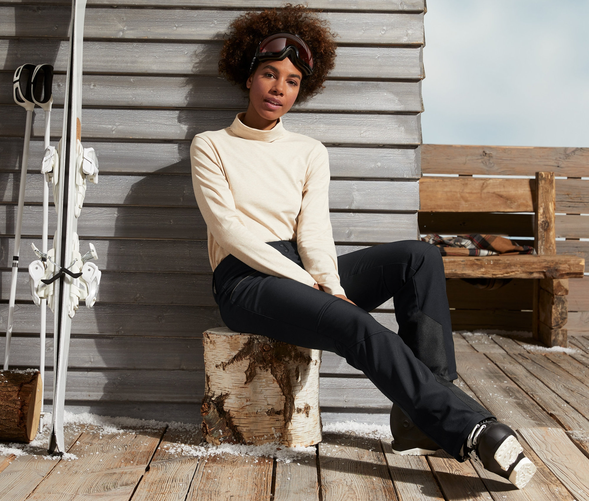 Femme assise sur un billot de bois en pantalon de ski élastique noir et chaussures de ski.