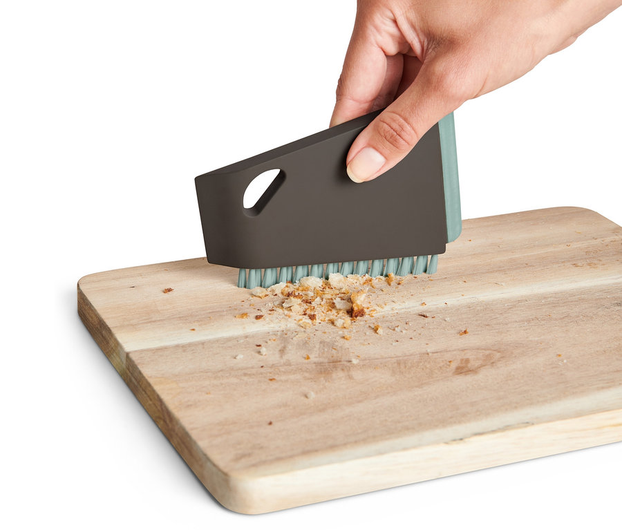 Une main utilise une brosse de cuisine universelle pour enlever les miettes d'une planche à découper en bois.