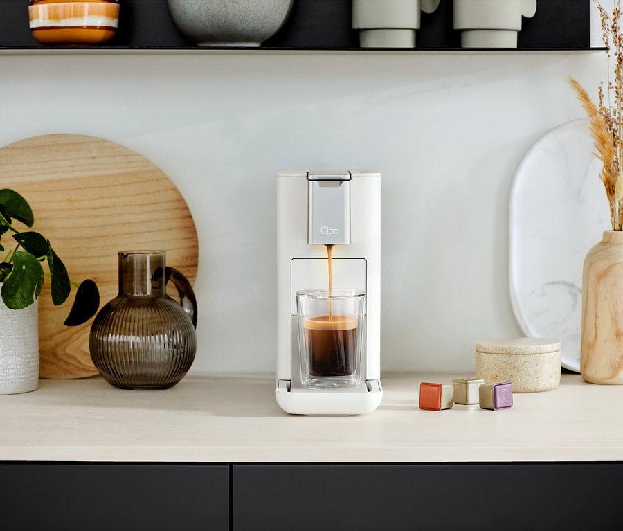 La machine à café Qbo ICONIC Dark Chrome et Qbo ESSENTIAL Bright Sand remplit un verre de café. Des capsules sont à côté.
