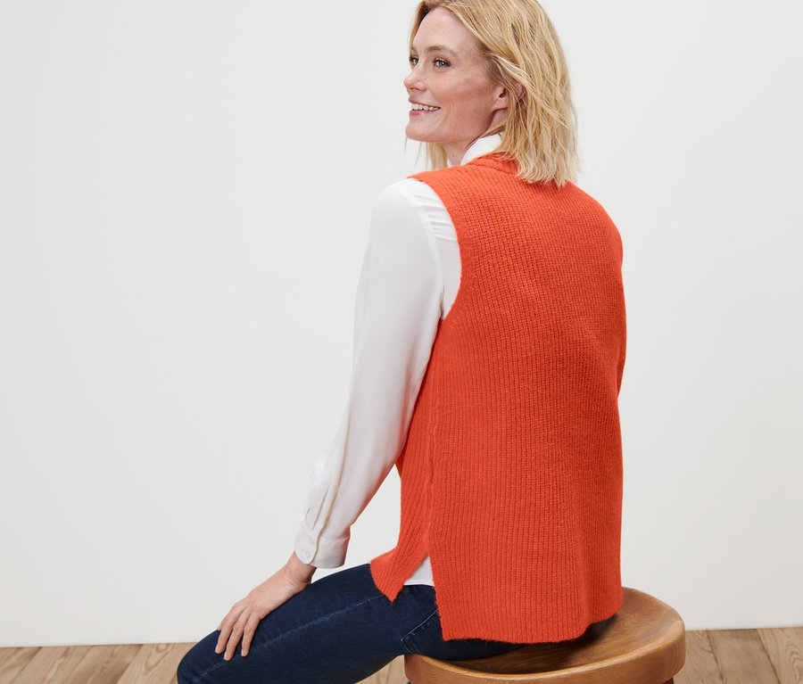 Femme assise sur un tabouret portant un pull sans manches en maille orange.