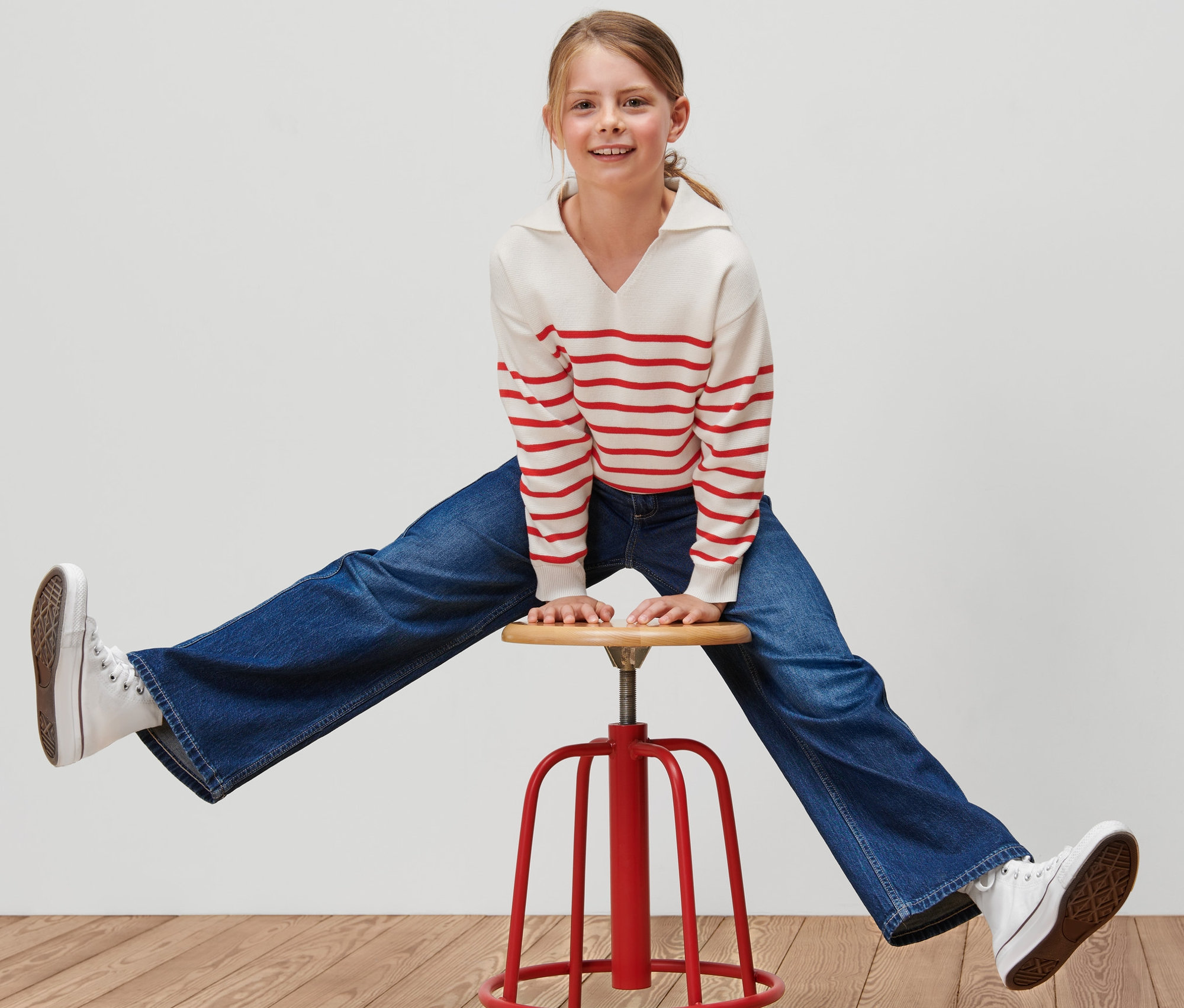 Une fille assise à califourchon sur une chaise rouge porte un pull en tricot pour enfant à rayures rouges, un jean bleu et des baskets blanches.