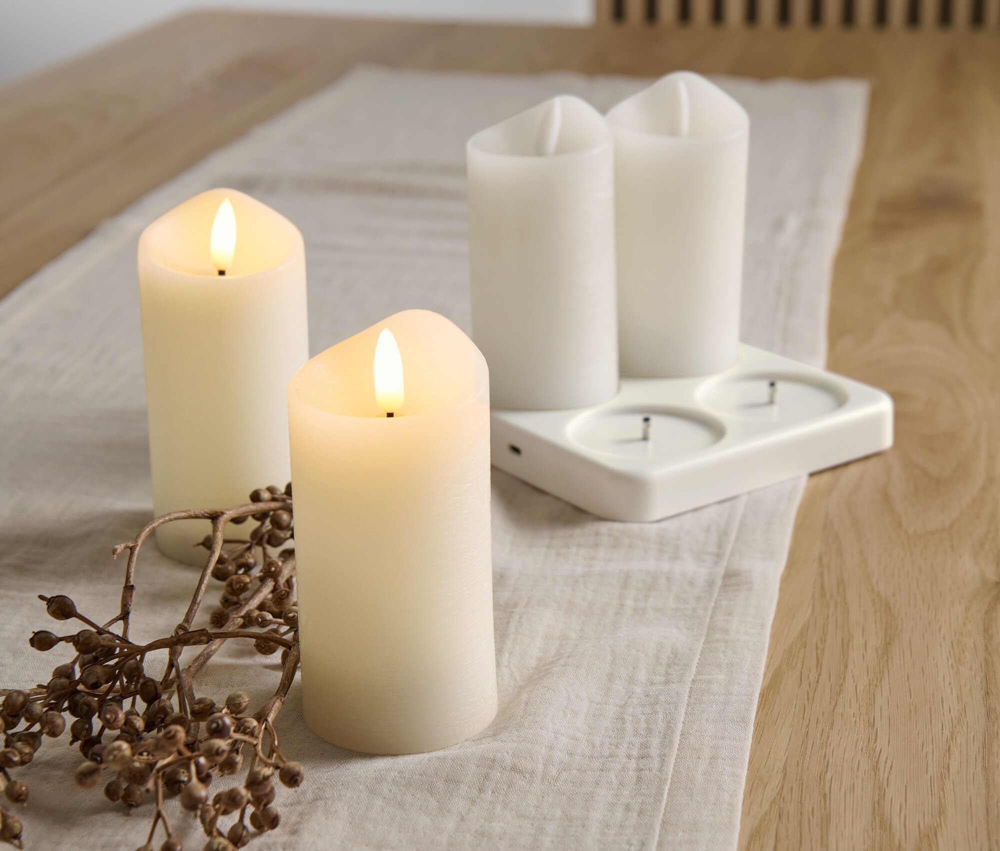 Sur une table, 4 bougies en cire véritable à LED sur batterie avec station de recharge et une branche avec des baies.