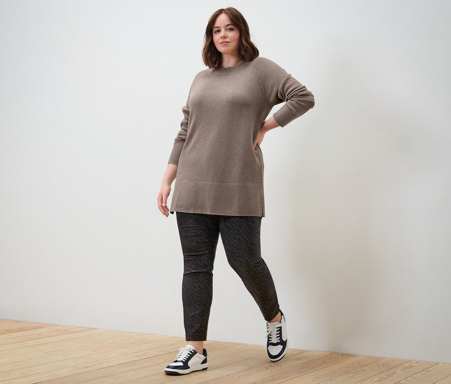 Une femme pose, portant un long pull en maille taupe et un pantalon en bengaline.