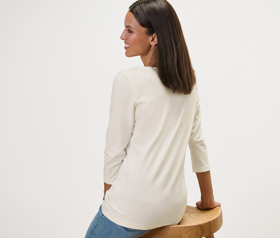 Une femme assise de côté sur un tabouret en bois porte un t-shirt blanc à manches 3/4.