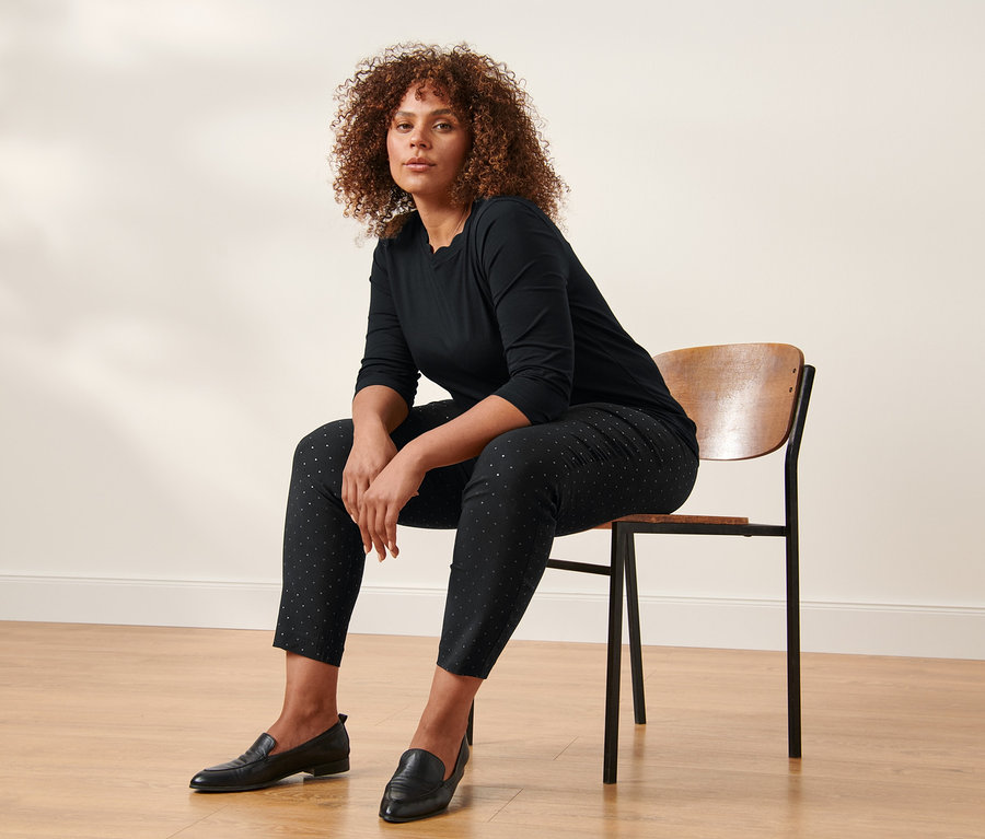 Une femme est assise sur une chaise et porte un t-shirt noir à manches 3/4 et un pantalon stretch à pois.