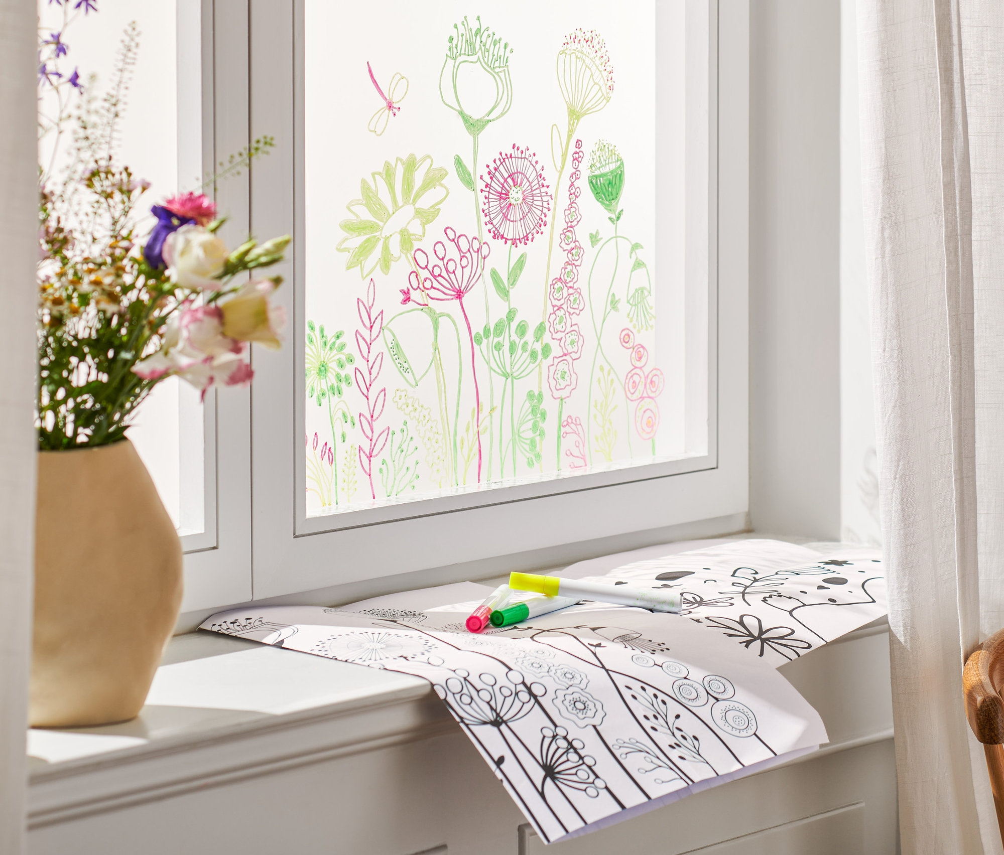 Des fleurs dans un vase sont posées sur un rebord de fenêtre, la fenêtre est peinte et sur le rebord se trouvent des peintures pour vitres pour Pâques et des marqueurs.