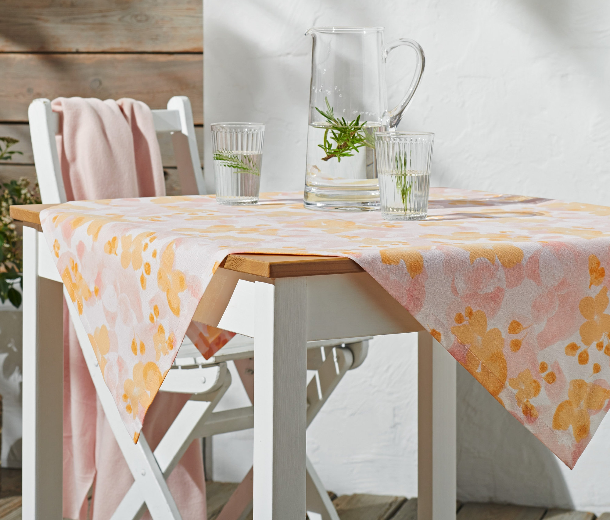 Sur une table de jardin blanche, une nappe d’extérieur moyenne d’environ 90 x 90 cm avec un motif floral orange. Sur la table, une carafe en verre et deux verres d’eau.