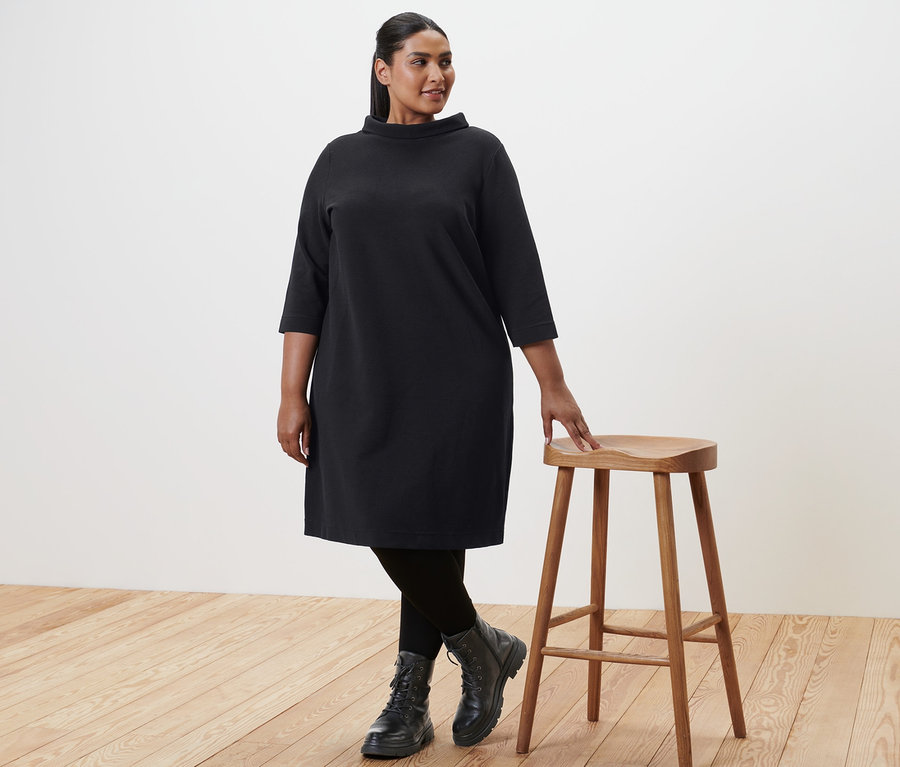 Femme portant des bottines en cuir noires et une robe sweat manches 3/4, debout à côté d'un tabouret en bois.