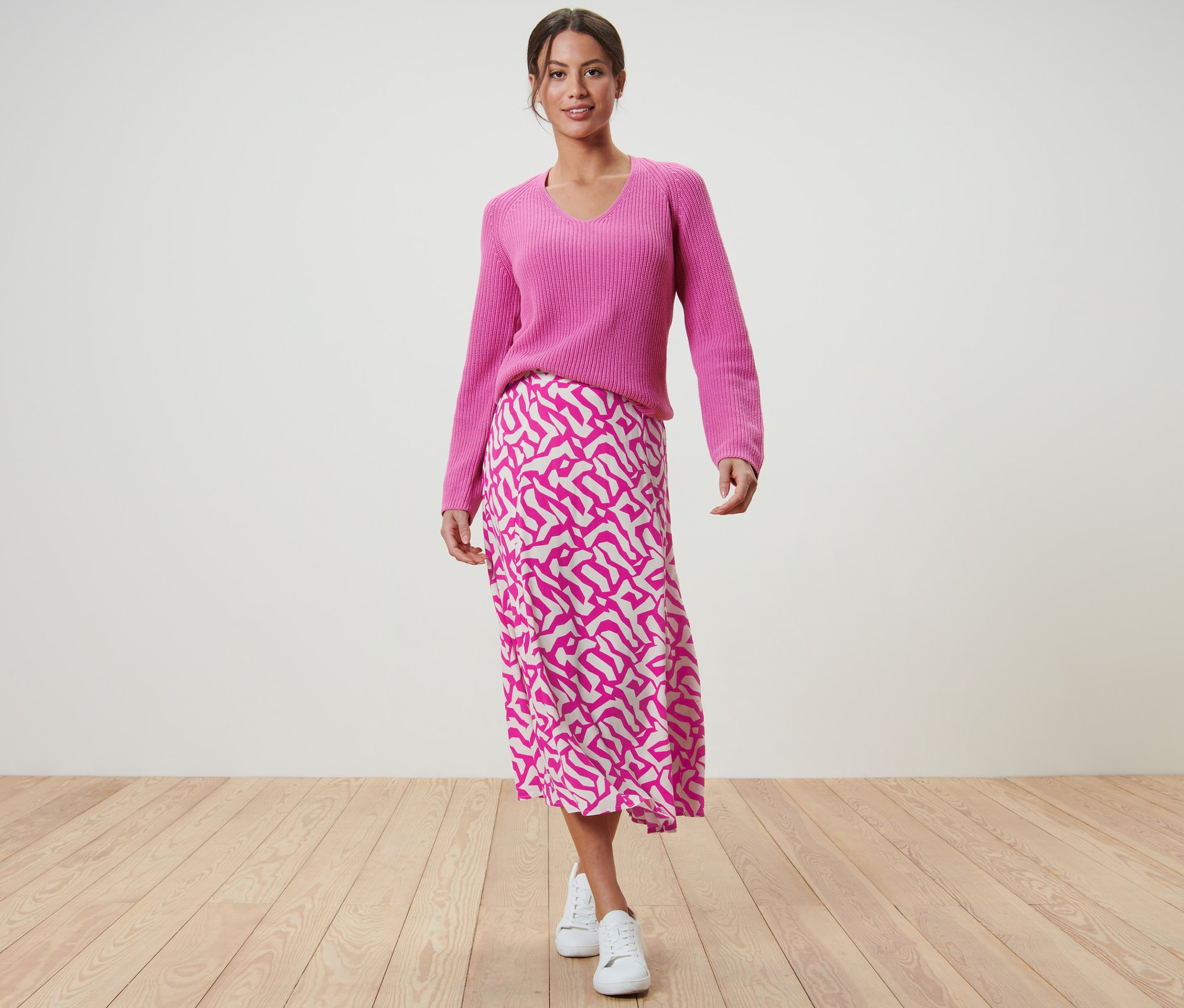 Une femme pose en pull rose et jupe midi imprimée rose vif.