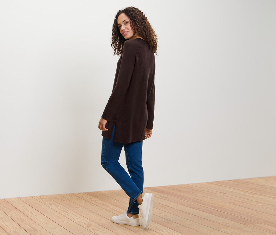 Une femme regarde par-dessus son épaule et pose, portant un long pull en maille marron foncé, un jean et des baskets.
