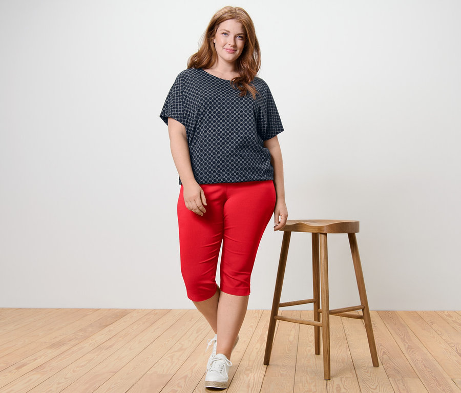 Une femme se tient à côté d'un tabouret en bois, portant une blouse imprimée et un pantalon 3/4 stretch rouge.