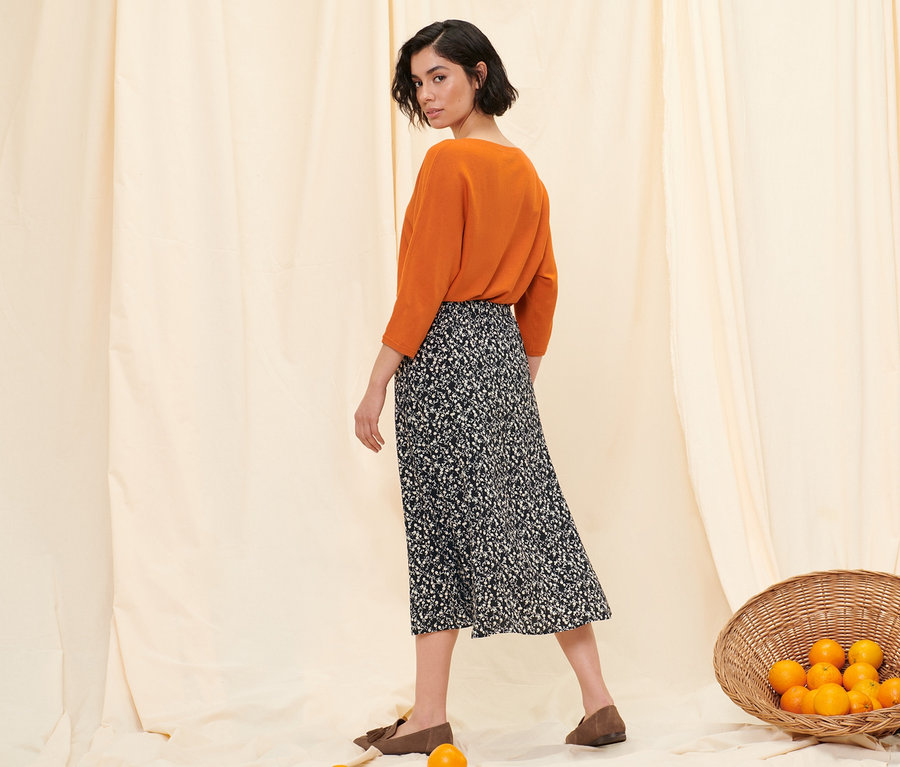 Femme posant en pull en maille fine orange, jupe midi florale et mocassins en cuir velours.