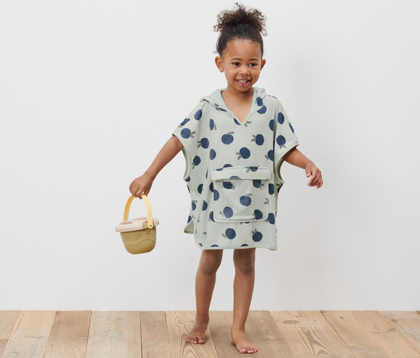 Fillette souriante portant un poncho en éponge pour tout-petits et tenant un seau jaune.