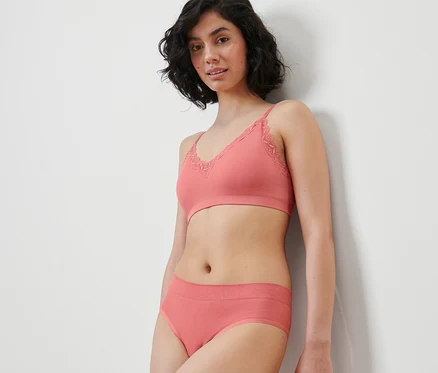 Une femme appuyée contre un mur porte un soutien-gorge corail et 2 slips brésiliens Seamless.