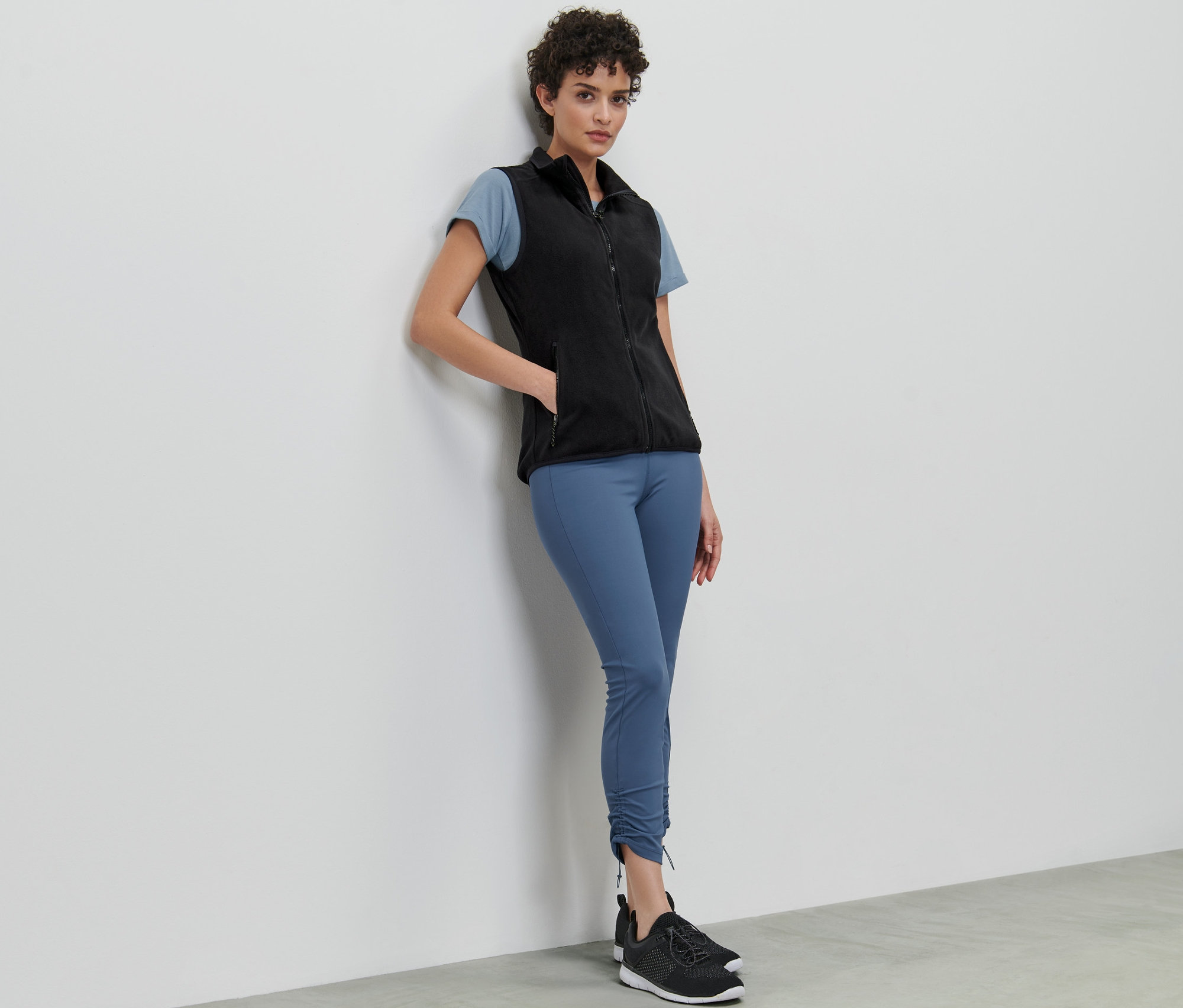 Une femme appuyée contre un mur porte un gilet sans manches en polaire, un t-shirt et un legging.