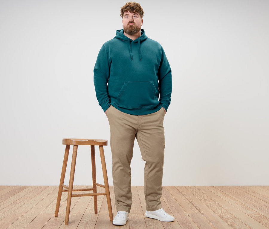 Un homme debout, les mains dans les poches. Il porte un sweat-shirt à capuche bleu pétrole et un pantalon chino beige.