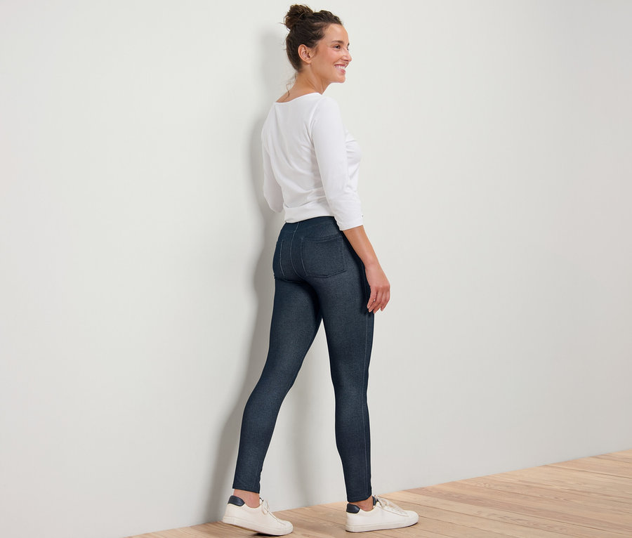 Une femme portant un legging aspect jean, un haut blanc et des baskets, se tient debout près d'un mur.