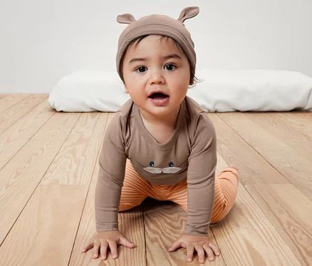 Ensemble pour bébé : Un bébé rampe sur un plancher en bois, vêtu d’un body brun imprimé et d’un pantalon rayé orange. Bonnet brun à oreilles.