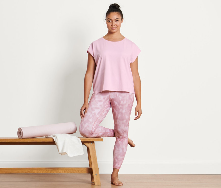 Une femme pose sur un banc portant un t-shirt de sport rose clair et un legging de sport réversible rose.