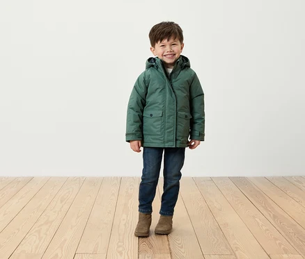 Un garçon souriant se tient sur un plancher en bois, portant une veste verte, un jean et des bottes marron.