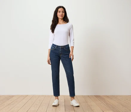 Une femme se tient debout et porte un t-shirt blanc à manches longues, un jean bleu foncé et des baskets blanches.