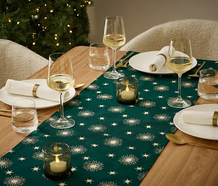 Table dressée avec un chemin de table vert foncé à motifs d'étoiles, des assiettes avec des serviettes, des verres à vin et à eau, et des bougeoirs avec des bougies allumées.