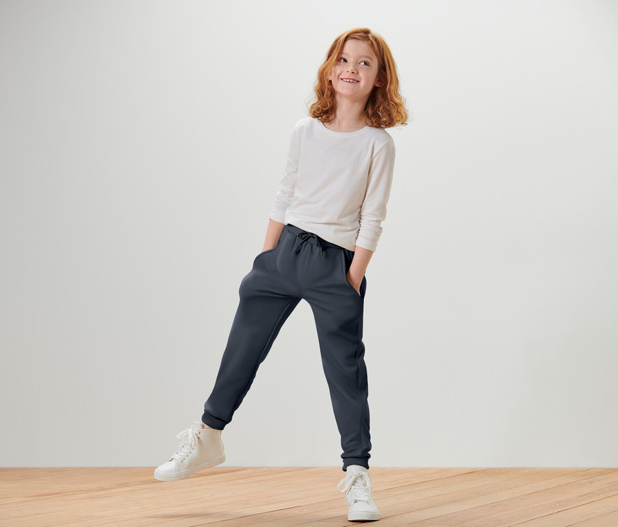 Fillette rousse portant un t-shirt blanc à manches longues et un pantalon de jogging bleu foncé, debout sur une jambe et souriant.