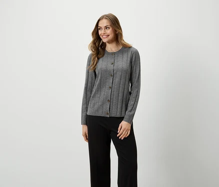 Une femme aux cheveux longs pose en cardigan gris boutonné et pantalon noir.